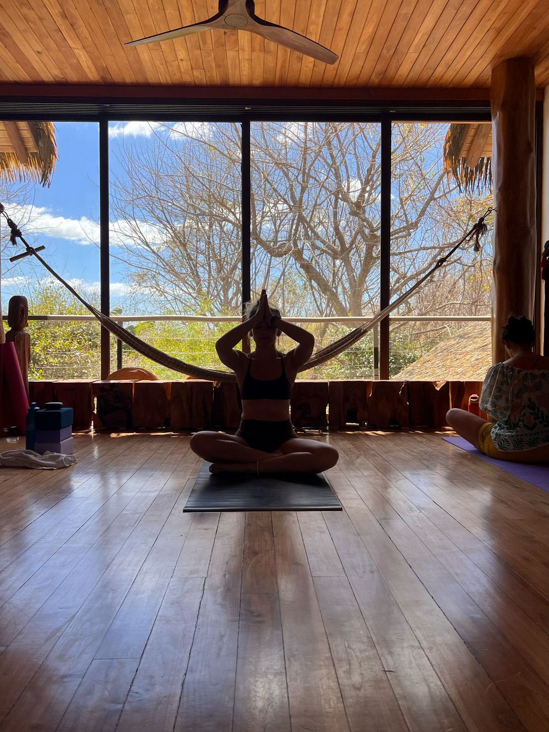 A woman seated in meditation on the villa&rsquo;s open-air yoga deck, jungle framed in the windows behind her